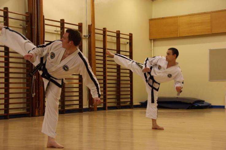 Mr Chan and Mr Lopez performing high reverse turning kick in a school hall. 