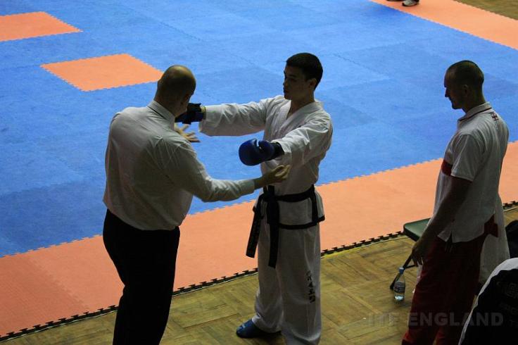 black belt standing next to a competition mat while his coach watches the umpire check his pads and feet. 