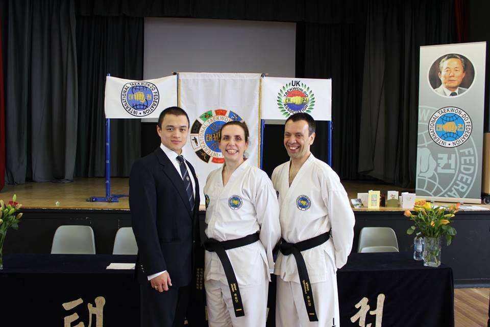 Post grading promotion photograph showing two new black belts and their instructor in front of ITF and UKTA banners and the grading table