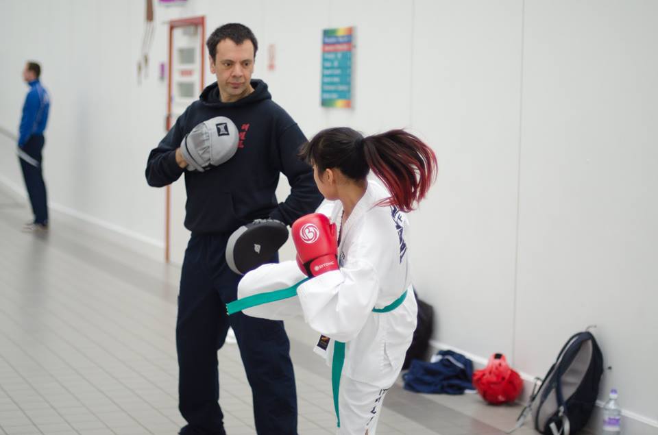 Black belt Alex Aquino holding hand pads for a green belt female student in Dobok and pads while she is kicking