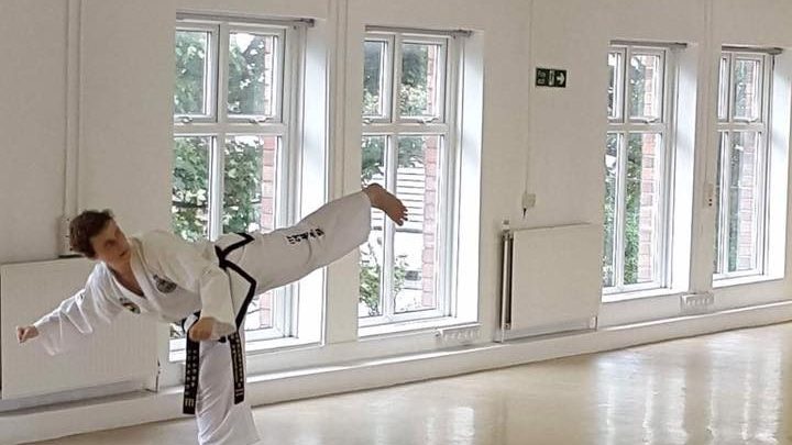 Eddie Dobson in Dobok performing a high section back kick with guarding block in naturally lit dance studio