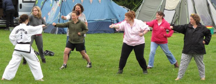 beginner taekwon-do lesson for the women's institute in a field. showing a black belt demonstrating walking stance punch to six women doing the same and smilin