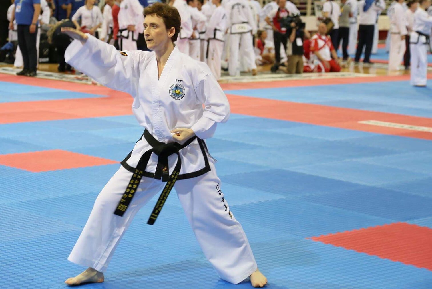 Eddie Dobson IV dan performing a high section knife hand strike in walking stance at a european championships. You can see red and blue competition mats and other competitors in the background