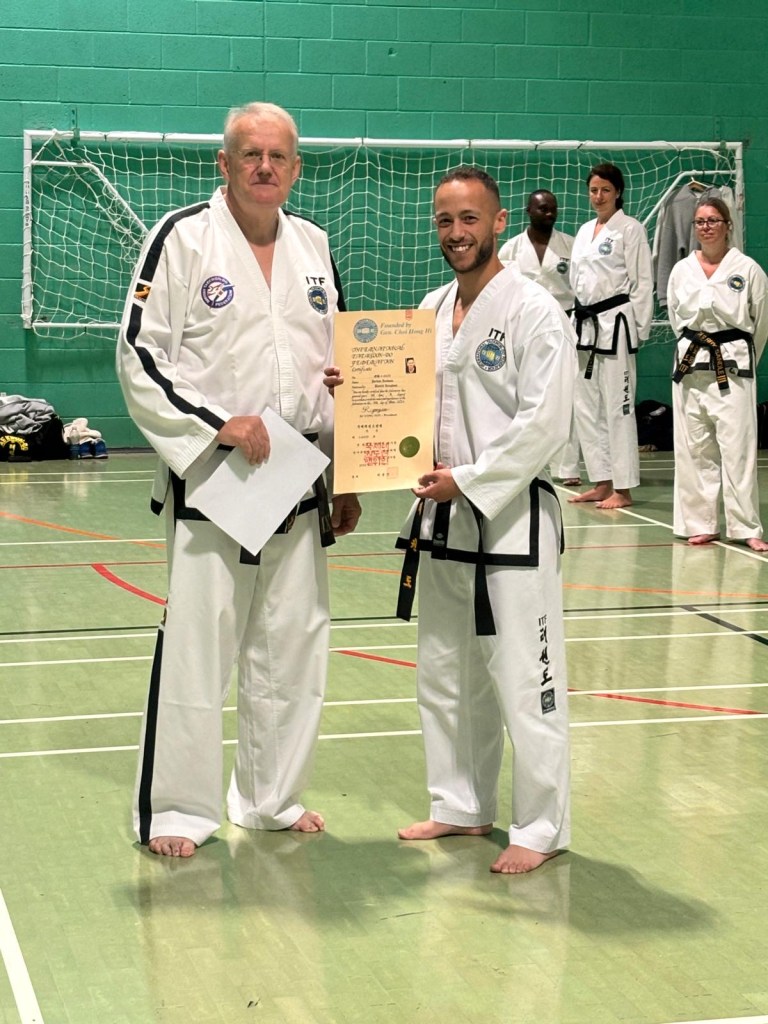 Mr Jordan Jackson receiving his ITF 1st dan black belt certificate from Grandmaster Wayne Brown IX Dan at a British Taekwon Do federation seminar and grading event. Picture shows both smiling, while Jordan holds his certificate up for the camera. Several black belts stand in the background. They are in a community sports centre. 